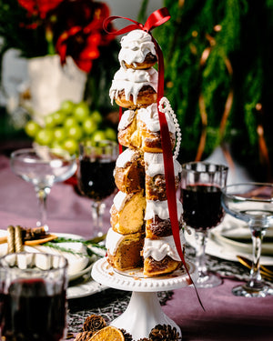 Tower of holiday bakery treats cake with white icing and red ribbon on a white stand at a festive table
