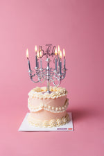 Gateau Gourmet Cafe Lumiere birthday cake with elegant white frosting and lit candles on pink background