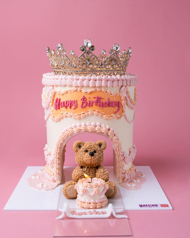 Princess crown cake for girls with pink and white frosting, a gold crown topper, and teddy bear decoration