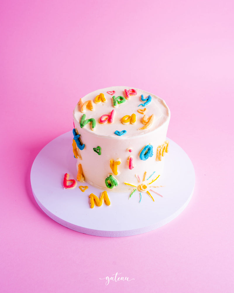 Scrabbled birthday mini cake decorated with colorful letters and cheerful designs on a pink background