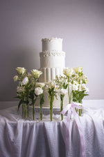 White wedding cake with flowers by Gateau Gourmet Cafe on a white draped table with floral decorations